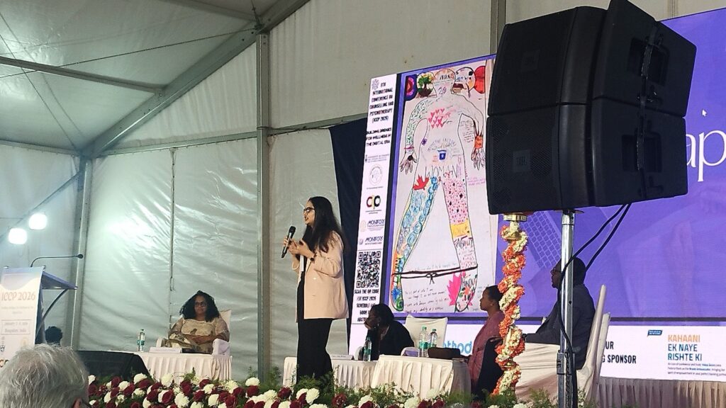Tushti Hari presenting her body map on stage at ICCP 2026 in Bangalore, the map displayed on the screen behind her.