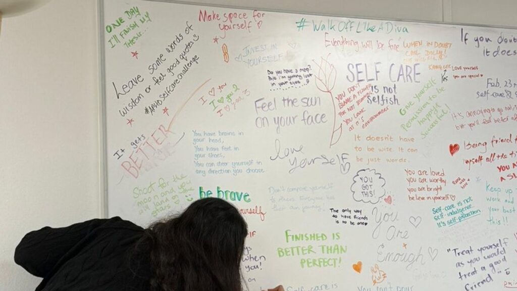 A whiteboard covered in handwritten self-care messages in many colours, including treat yourself as you would treat a good friend, and self-care is not self-indulgence it is self-protection, with someone adding another line.