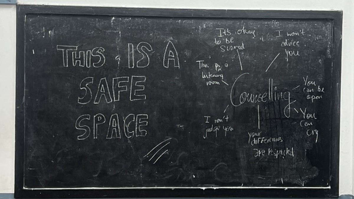 A classroom blackboard with THIS IS A SAFE SPACE written in chalk, surrounded by handwritten phrases about counselling: it is okay to be scared, I will not judge you, this is a listening room, you can cry.